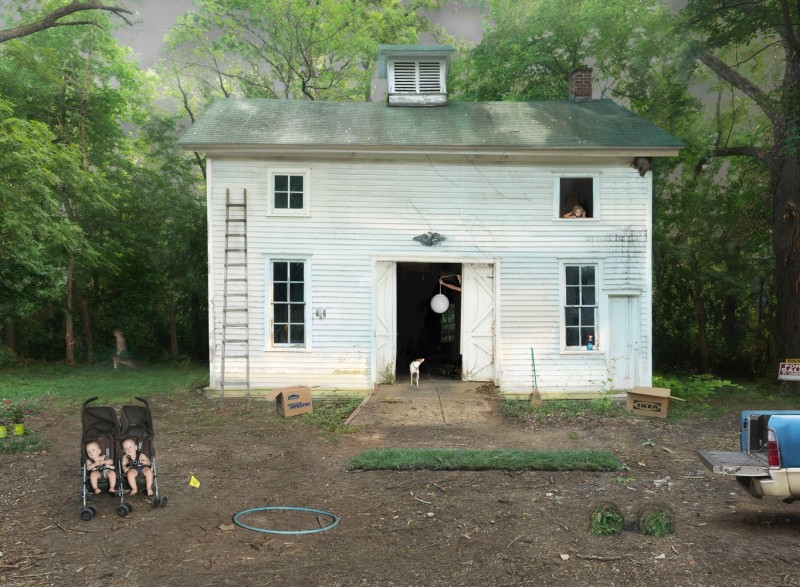 An old house is shown with the door wide open, a dog in the doorway looking up at a child in the upstairs window, and two babies in a twin stroller in this color photograph.