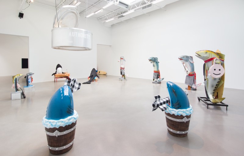 Installation view of WHAT IF IT BARKS? at Petzel in 2018. The image features eight plastic fish and two fabric killer whales in a large circle with a giant aluminum cat food can hanging from the ceiling.