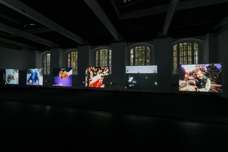 Six videos by price projected on side-by-side screens of the same size. The lights are off but the arched windows behind show the green silhouette of trees in the day time.
