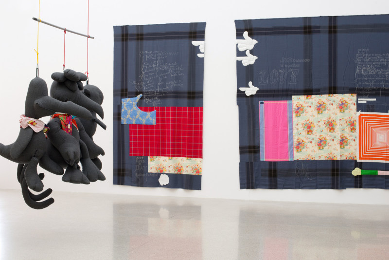 Installation view of von Bonin's show at MUMOK featuring three large hanging stuffed birds, each wearing colorful scarves. Two patch-work cloth &quot;paintings&quot; hang in the background.