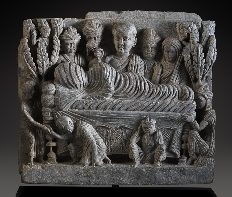 This relief shows the parinirvana of the Buddha in a rather unusual manner.  The teacher, on his death bed, is depicted at the center of the relief.  His head and body are completely swathed in cloth, and his face is not visible.