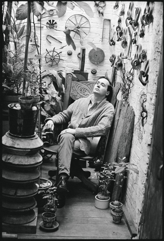 A black and white photograph of Robert Indiana seated in the plant room of his Coenties Slip studio. Wheels and other hardware are seen on the back wall