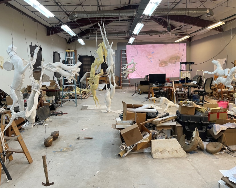 an artist studio with sculptures of animals hanging from the ceiling, an assortment of boxes, art materials, and chairs and desk off to the right with a video projection on a portion of the back wall