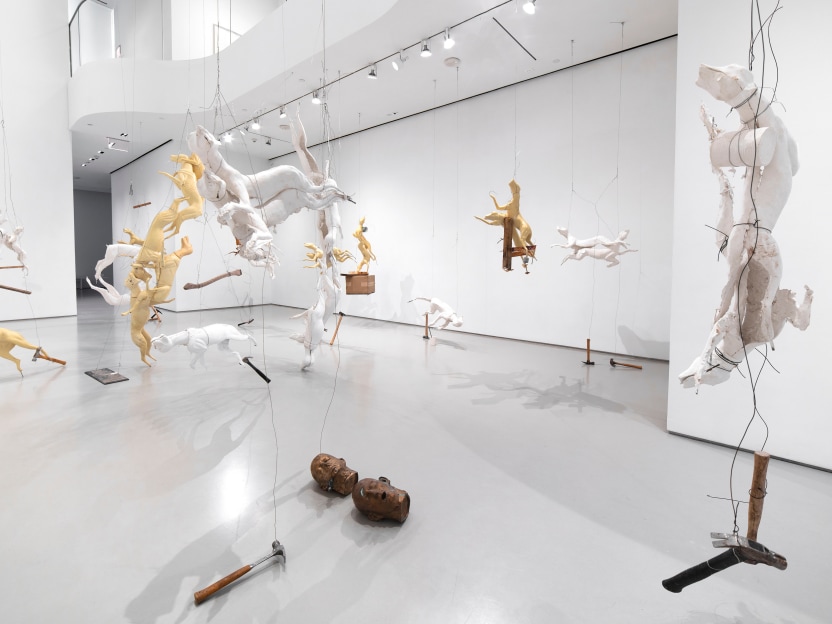 gallery exhibition with 17 sculptures suspended from the ceiling. the sculptures are composed of taxidermy forms of foxes, coyotes, hammers, and other found materials