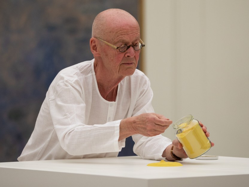 The artist, Wolfgang Laib, spoons yellow pollen from a jar onto a white pedestal, creating a mound of pollen.