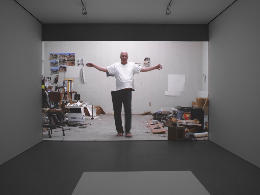 video projection onto a white wall of an older man with arms stretched out walking in a straight line in art studio