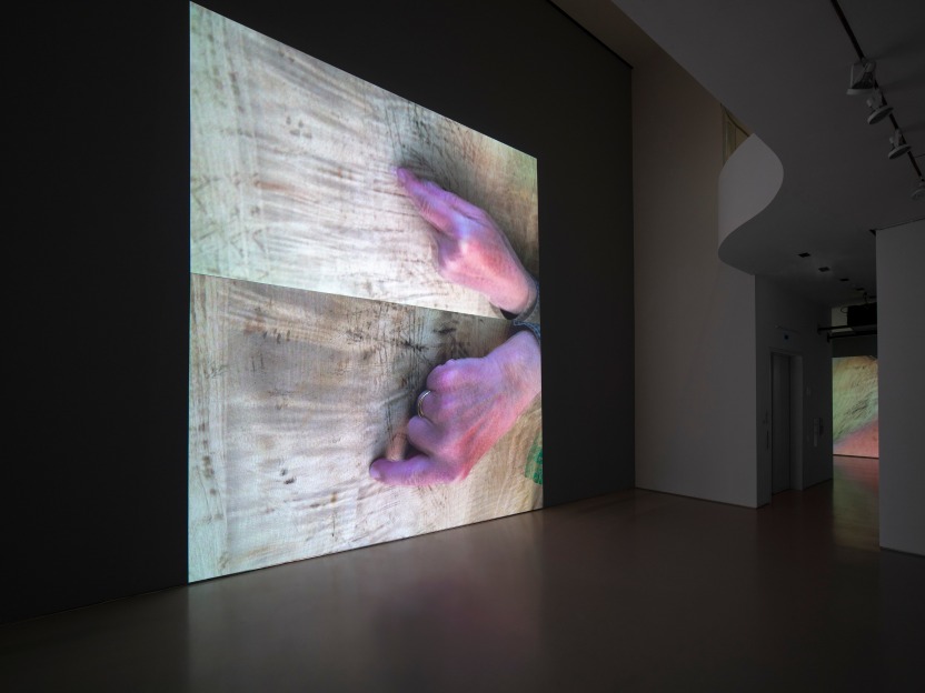installation view of two video projections on gallery wall depicting the artist's hands on a wooden table