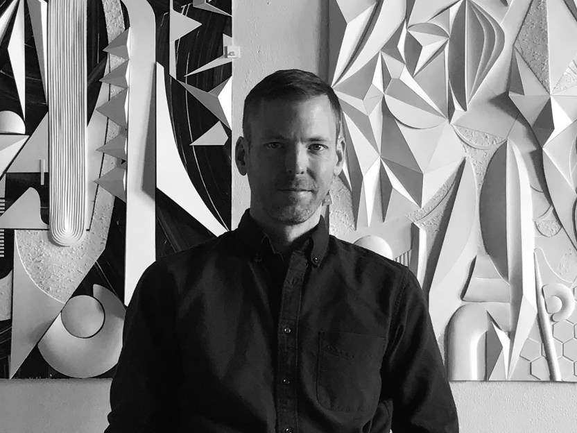 black and white portrait of artist Jim Gaylord in front of two abstract artworks hung on the wall behind him