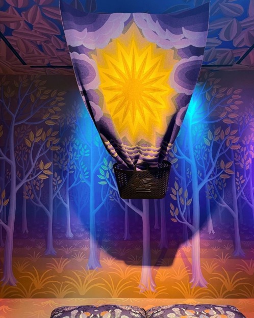 Miniature hot air balloon with a bright yellow sun,  in front of a purple tapestry with bare trees and two grey pillows directly underneath