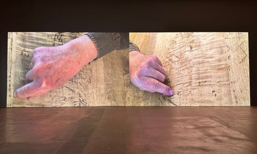 two large-scale projections side by side each showing  an age hand with index fingers extended on a scratched wooden surface