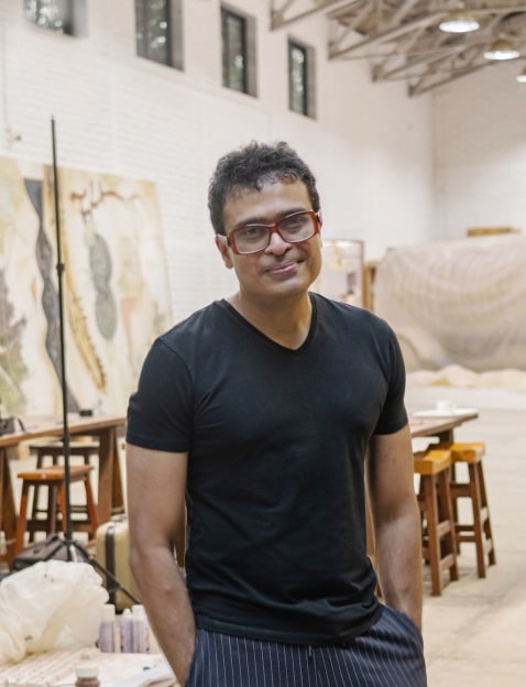 A man wearing red glasses stands with his hands in his pockets in a white artist studio