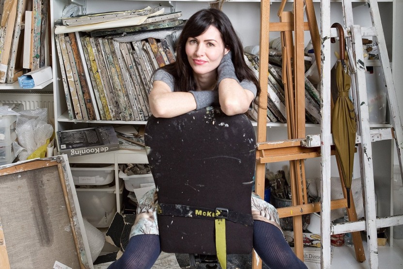 Dark haired woman sitting on a backwards chair in an artist's studio surrounded by paintings, easels and an olive green umbrella. 