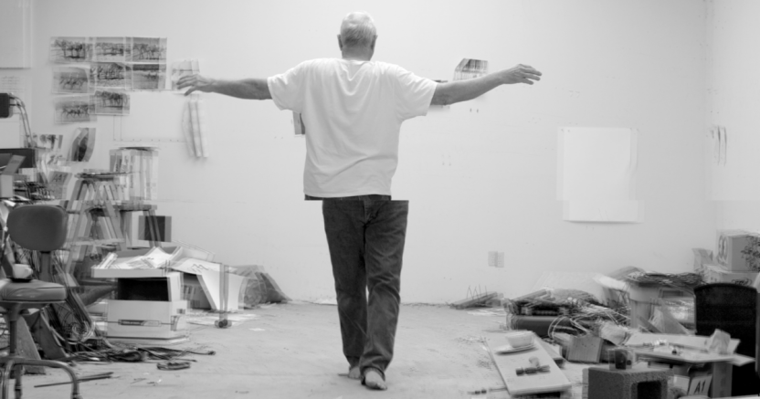 A man wearing a white tshirt and blue jeans steps toward the camera while his upper half faces away with his arms stretched out, in a cluttered artist's studio