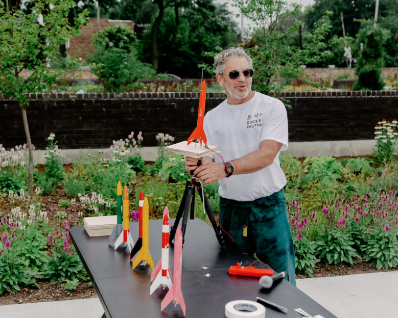 Tom Sachs stands at an outdoor table with colorful model rockets in a garden-like setting