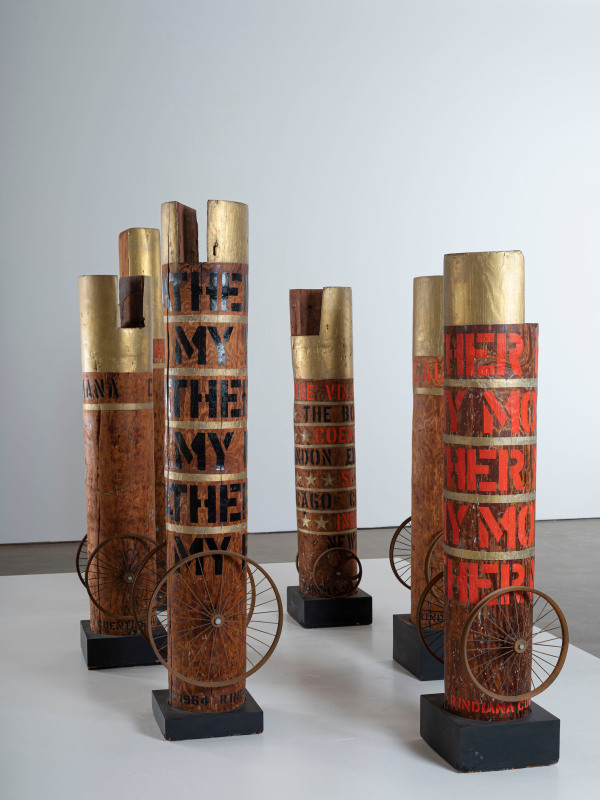 Installation view of six of Robert Indiana's columns. Each of the columns has two wheels attached to the bottoms sides, and a band of gold paint around the top of the columns