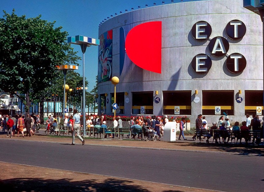 World's Fair installation view of Indiana's EAT, an X-shaped polychrome and stainless steel aluminum sculpture consisting of five black circles spelling the word EAT twice diagonally in white letters with light bulbs. Two circles contain the letter E, on circle the letter A, and two circles the letter T.