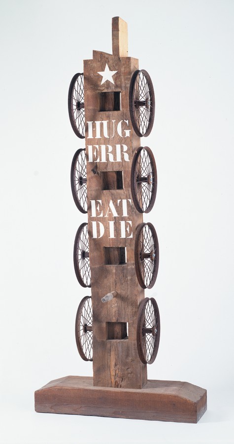A sculpture consisting of a wooden beam with a haunched tenon on a wooden base. Four iron wheels run down the left and right sides of the sculpture. At the front top is a white star, below it the words "Hug," "Err," "Eat," and "Die" are painted in white stenciled letters.