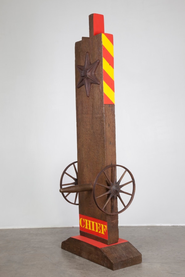 A 63 13/16 by 23 1/8 by 9 1/8 inch painted bronze sculpture of a beam with a haunched tenon, and on a base. The sculpture's title, "Chief," is painted in yellow stenciled letters against a red rectangular ground across the bottom of the work. Above the title a wheel is attached to the right and left sides of the sculpture, and a peg has been placed in the front of the sculpture, in between the wheels. At the top of the sculpture is a six pointed star. The top third of the right and sides of the sculpture are painted with diagonal red and yellow stripes, and the sides left and right sides of the tenon are painted red.
