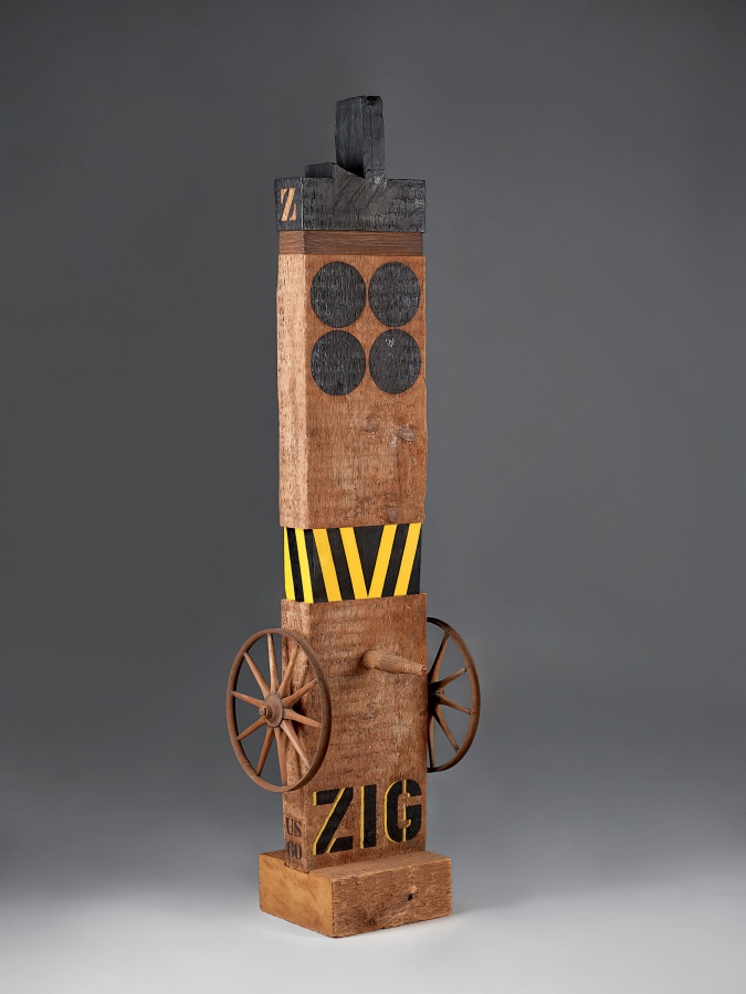 A sculpture consisting of a wooden beam with a haunched tenon on a wooden base. The work's title, "Zig," is painted in black stenciled letters across the bottom of the work. Above the title, affixed on the left and right sides of the sculpture, are metal wheels. A wooden peg has been affixed to the front of the sculpture, in between the top rims of the wheels. A few inches above the peg is a band of yellow and black vertical stripes. The tenon and top of the sculpture have been painted black. Below this is a band of thin metal wires, and below that two rows of two black circles.