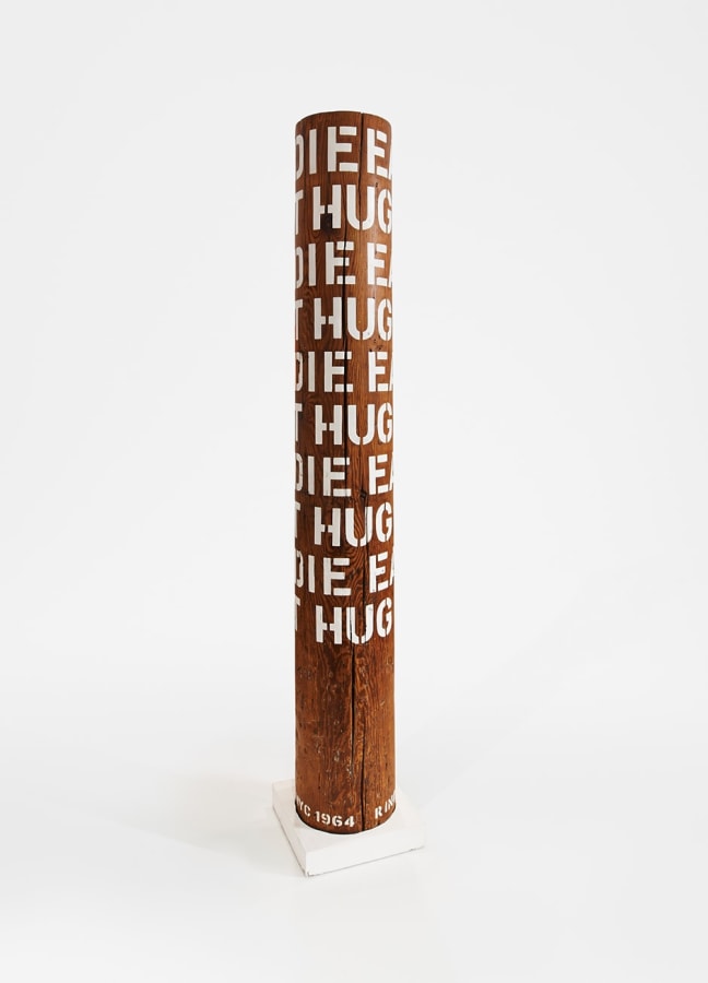 A column with ten rows of the words eat, hug, and die in gesso stenciled letters wrapping around the upper three quarters of the sculpture. 