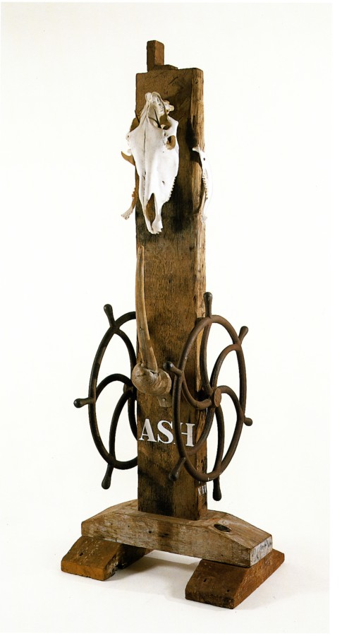 A sculpture consisting of a wooden beam with a haunched tenon on a wooden base atop two forward facing wood risers. The sculpture's title, "Ash," is painted in white letters towards the bottom front of the sculpture, and a long wooden perpendicular peg has been affixed above the title. To the left and right, affixed to the sides of the sculpture, are iron wheels. An animal skull has been affixed to the top front of the sculpture.