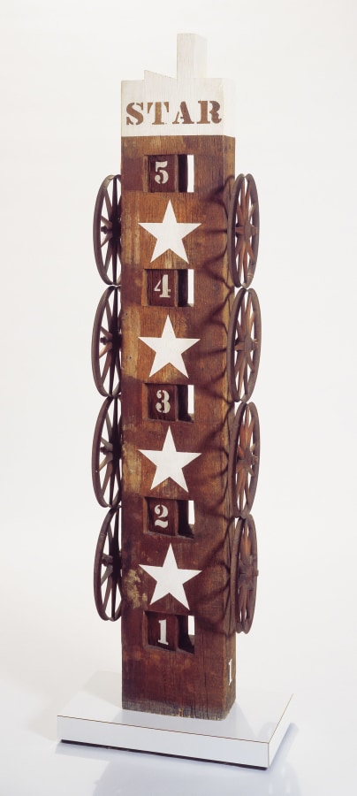 A sculpture consisting of a wooden beam with a tenon on a wooden base. Four iron wheels have been affixed to both the right and left sides of the sculpture, a white star has been painted between each set up wheels. White numbers, from one through five, have been painted down the front, starting with the number 5 above the top star, with the numbers counting downwards, 4, 3, and 2 appearing between stars and 1 below the bottom star. The top of the sculpture, above the number five, has been painted white, and contains the work's stenciled title, "Star."