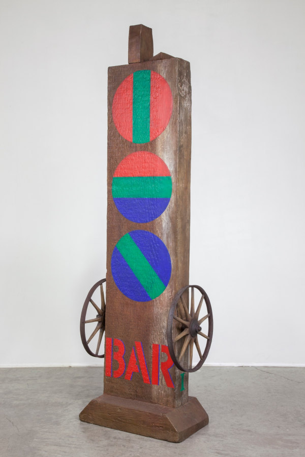 A 58 5/8 by 15 by 12 5/8 inch painted bronze sculpture consisting of a beam with a haunched tenon, on a base. The sculpture's title, "Bar," is painted in red stenciled letters across the bottom front of the work. Occupying the space from few inches above the title to the top of the sculpture are three painted red circles. The top one is red with a green vertical stripe down the center, the middle one consists of red, green and blue horizontal fields, and the bottom one is blue with a diagonal green stripe down the middle. A wheel has been affixed to the right and left sides of the sculpture, just above the title.