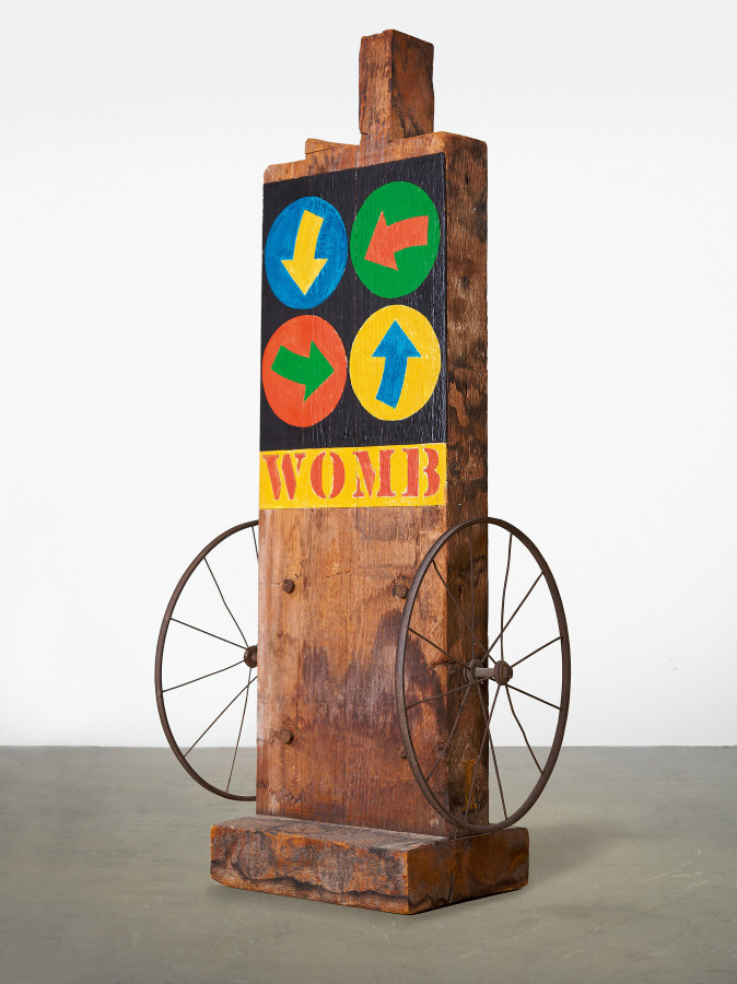 A sculpture of a wooden beam with a haunched tenon, on a wooden base. The bottom half of the sculpture has been left bare, and an iron wheel has been affixed to the right and left sides. At the bottom of the upper half is a yellow painted band, with the work's title, "Womb," painted in red stenciled letters. Above this are four circles containing an arrow, on a black ground. The top row contains a blue circle with a yellow arrow pointing downwards, and a green circle with a red arrow pointing to the left; and the bottom row contains a red circle with a green arrow pointing to the right, and a yellow circle with a blue arrow pointing upwards.