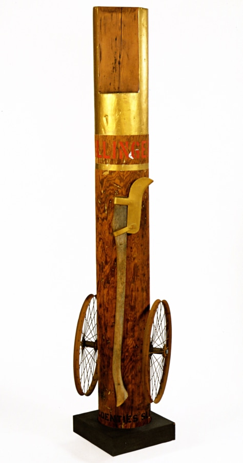 A column with an iron wheel affixed to the right and left lower sides, and standing on a wooden base. An iron axe is attached to the column. The top of the column is painted gold, and below is the work's title, "Dillinger," in red letters. Below the title a gold strip wraps around the column.