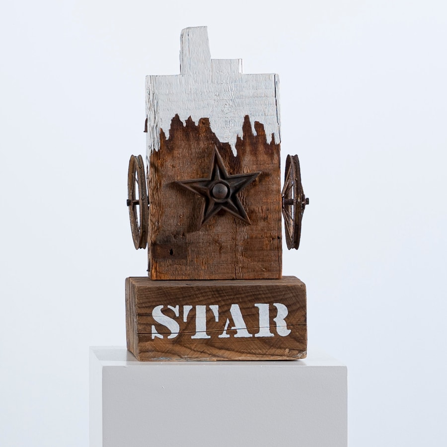 A sculpture consisting of fragment of a wooden beam with a haunched tenon, resting on a wooden base. The work's title, "Star," is painted in white stenciled letters across the front of the base. A metal star is affixed to the center front of the sculpture, and a small wheel has been affixed on both sides of the sculpture, to the left and right of the star. The top part of the sculpture has been painted white, with an uneven bottom edge, resembling dripping paint.