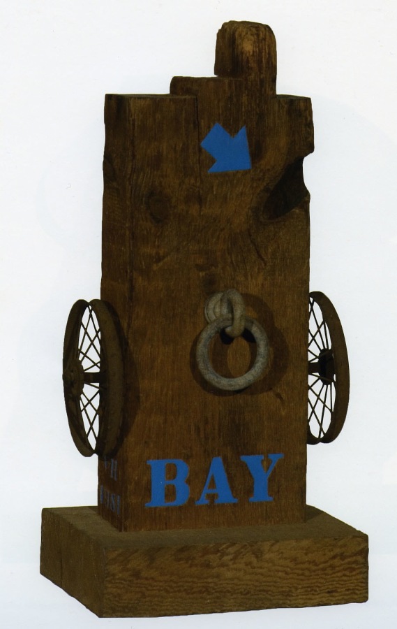 A sculpture consisting of a wooden beam with a haunched tenon. The work's title, Bay, is painted in blue letters across the lower front of the work, above the base. A small iron wheel is affixed on each side, and an iron ring on the front center. Above this is a painted blue arrow.