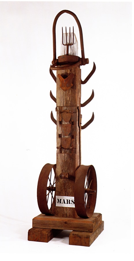 A sculpture consisting of a wooden beam atop a wooden base. The work's title, "Mars," appears in stenciled letters against a white ground across the front bottom of the sculpture. An iron wheel is affixed to both the bottom right and left sides of the sculpture. Three iron brush axes have been affixed to both the upper right and upper left sides of, and an iron pitchfork has been affixed at the top of the sculpture. 