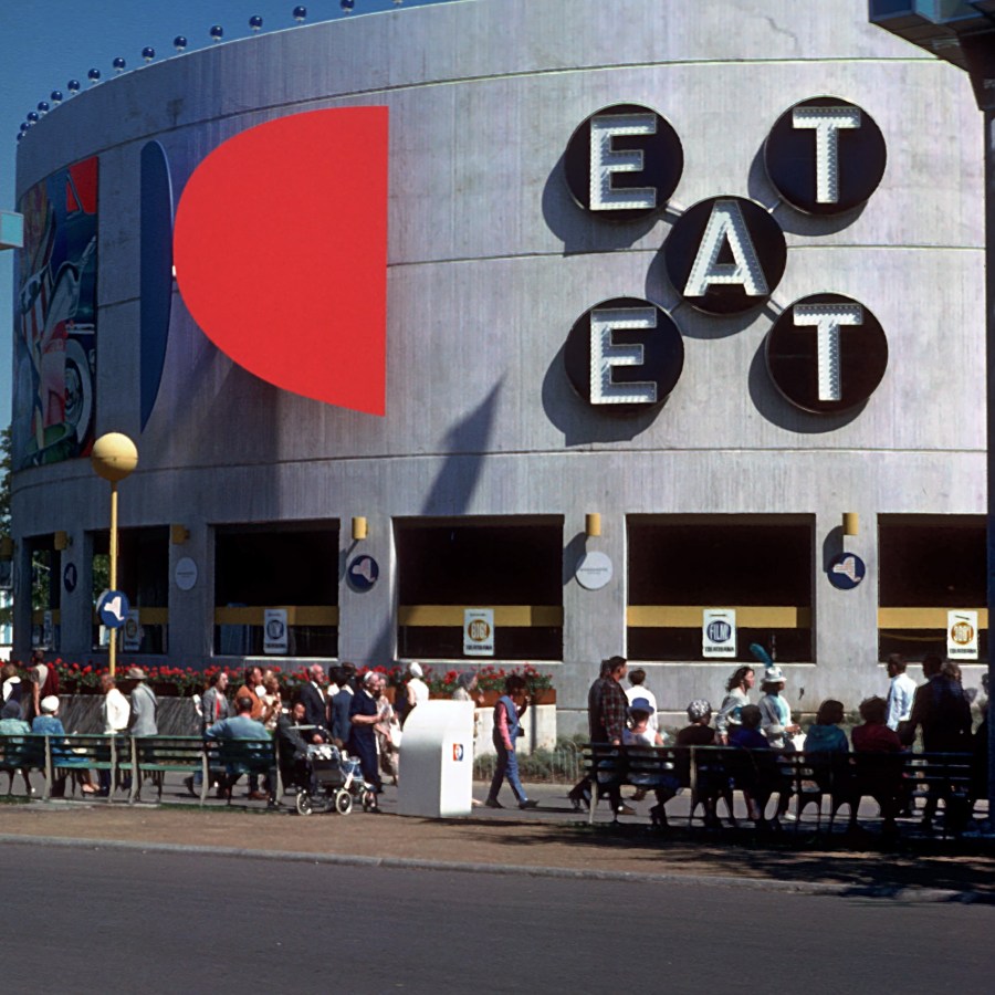 World's Fair installation view of Indiana's EAT, an X-shaped polychrome and stainless steel aluminum sculpture consisting of five black circles spelling the word EAT twice diagonally in white letters with light bulbs. Two circles contain the letter E, on circle the letter A, and two circles the letter T.