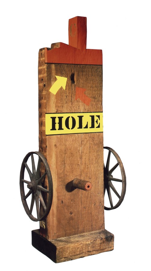 A sculpture consisting of a wooden beam with a haunched tenon on a wooden base. Iron and wooden wheels have been affixed to the lower right and left of the sculpture, in between the wheels, on the front of the sculpture, is a wooden peg. The work's title, Hole, appears above the wheels, painted in black stenciled letters against a yellow band of paint. Above the title are a yellow and a red arrow, each pointing towards a hole in the sculpture. The tenon and top front of the beam are painted red.