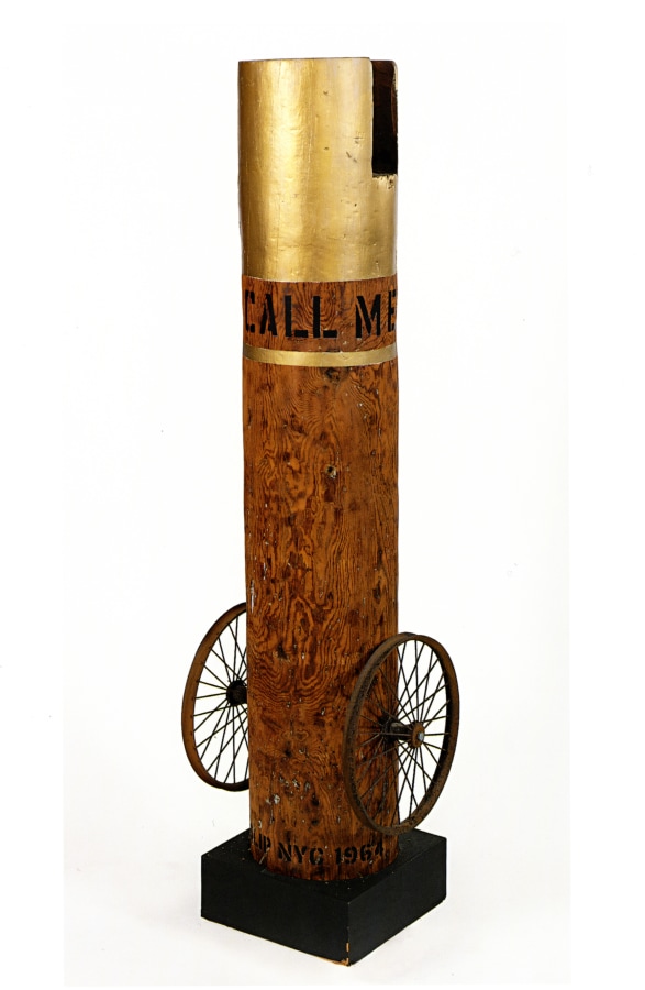 A 63 5/8 by 17 by 14 inch column with an iron wheel on the bottom left and right of the sculpture, and standing on a wooden base. The top of the column is gold, below, in black letters wrapping around the column, is its title, "Call Me Indiana." Below the title, wrapping around the column, is a gold stripe.