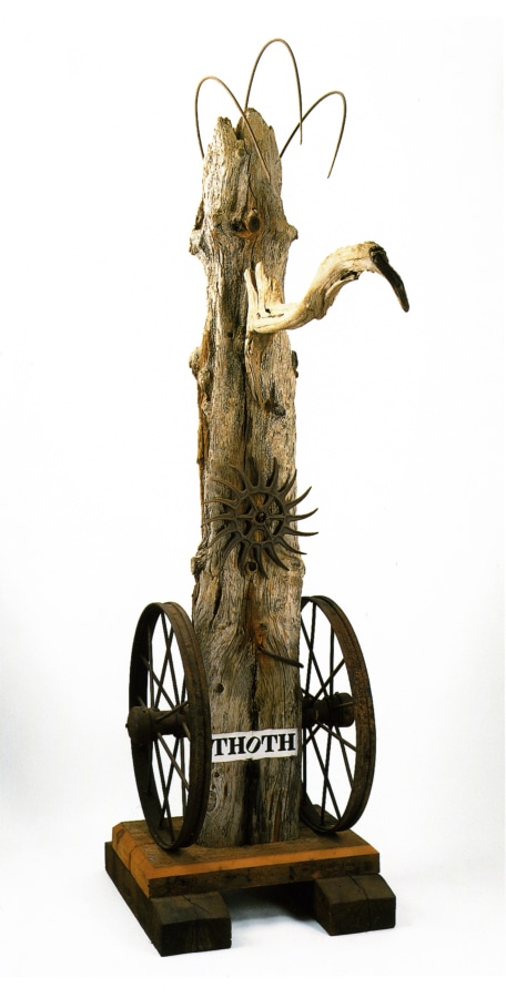 A weathered wood sculpture on a wooden base. The sculpture's title, "Thoth," is painted in black stenciled letters on a white ground across the front of the sculpture, near the base. An iron wheel has been attached to the sides of the sculpture to the left and right of the title. A long metal nail protrudes from the sculpture above the title, and above this is an old rotary tiller wheel. A piece of driftwood, resembling a long beak, has been attached to the front of the work near the top. 