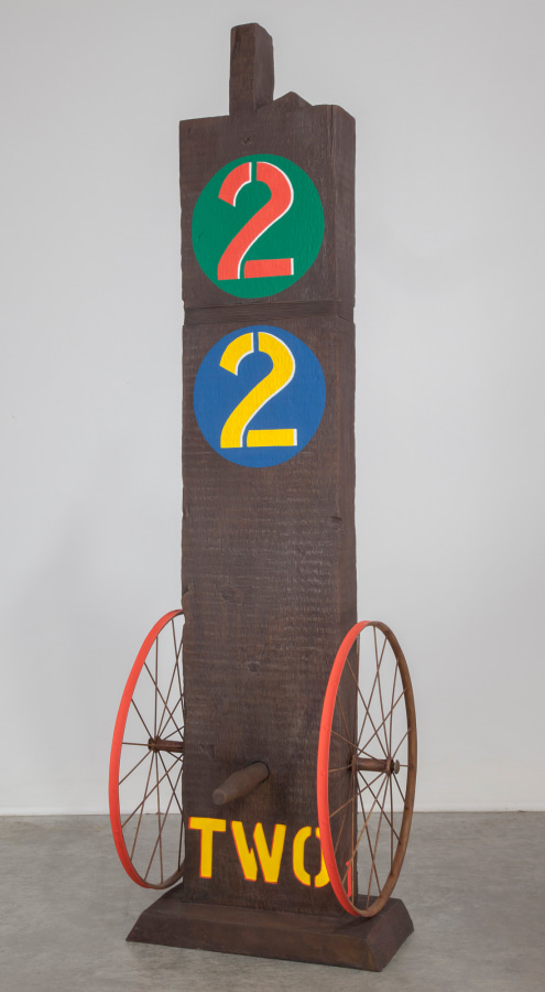 A 61 1/8 by 18 1/2 by 19 1/2 inch painted bronze sculpture of a beam with a haunched tenon, on a base. The work's title, "Two," is painted in yellow stenciled letters across the bottom front of the sculpture. A wheel is attached to the bottom left and right sides of the sculpture, and a peg has been affixed to the front of the sculpture, in between the wheels. At the top of the sculpture is a green circle with a red numeral two, and below that is a blue circle with a yellow numeral two.