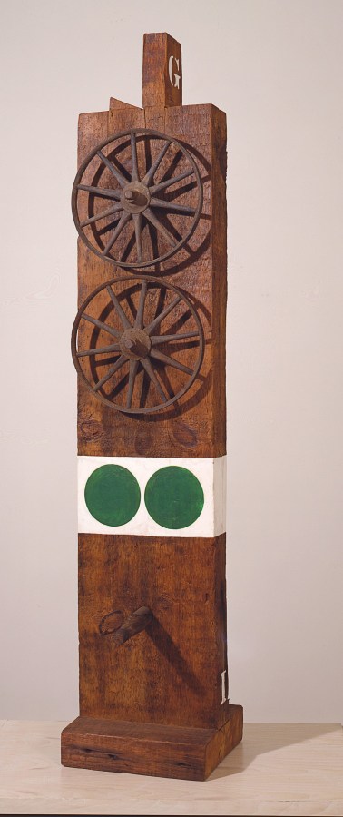 A wooden sculpture consisting of a wooden beam with a haunched tenon on a wooden base. On the top front of the sculpture are two iron and wooden wheels. Below the wheels is a white band of paint with two green circles. Halfway below the white band and the base a wooden peg protrudes from the sculpture.