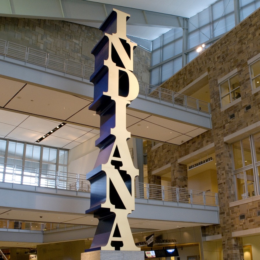 A polychrome aluminum sculpture spelling the word Indiana vertically, starting with the I on top and ending with the A on the bottom. The letters have gold faces and blue sides.