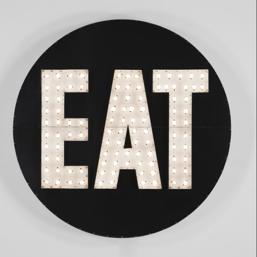 A 78 by 78 by 7 inch aluminum and stainless steel black circular sculpture of, with the word EAT in white with light bulbs.