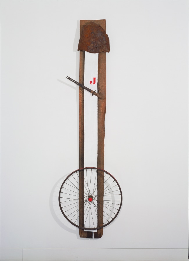 A hanging construction consisting of a long rectangular wooden plank. Mounted on top of this is a thinner white gessoed board. Towards the bottom front of the sculpture is a metal and wood wheel. A two-pronged tool with a broken wooden handle has been affixed diagonally in the lower upper third of the work. Above this is a painted red letter J. At the top of the white board is a flattened semi-circular piece of rusted metal.