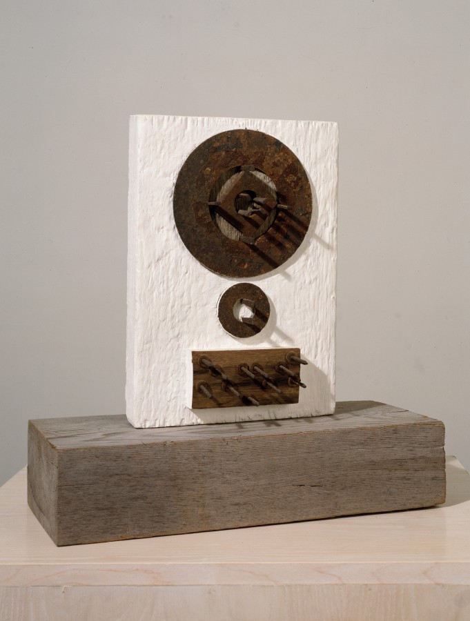 A sculpture constructed from a gessoed fragment of a wooden plank. Three found, rusted metal elements are held in place on down the front of the work with masonry nails, which face outwards towards the viewer. The top element is circular and the largest, the middle element is also circular, and the bottom element is rectangular. The work stands on a block of wood.