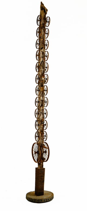A sculpture consisting of a severely weathered log standing on a circular base. Twelve wheels are affixed to each side of the sculpture, all the same size except for the first set, which is larger. In between the two lower wheels is a peg.