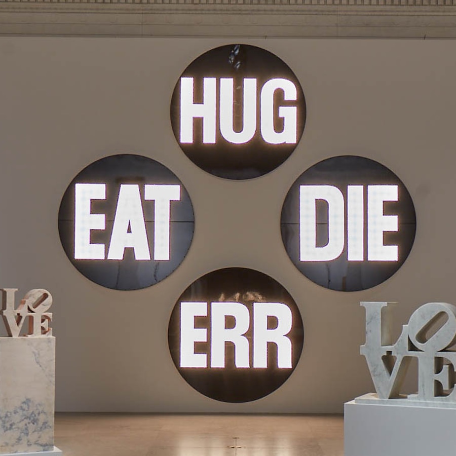 A sculpture consisting of four circular components. Each contains a different three letter word with lightbulbs that flash on and off. The words are eat, die, hug, and err.