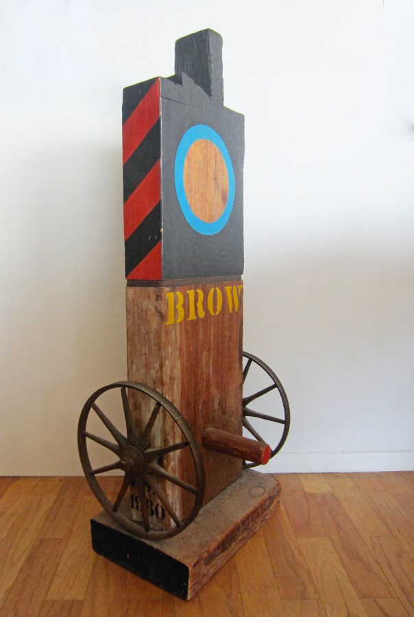 A sculpture consisting of a wooden beam with a haunched tenon on a wooden base. The top half of the sculpture has been painted black, with the exception of a blue containing an exposed wood circle in the top front center, and the top left and right sides, which have been painted with diagonal red and black stripes. The bottom half of the sculpture has been left unpainted, except for the title, "Brow," painted in yellow stenciled letters in the middle of the work. An iron wheel has been affixed to the lower left and right sides of the work, and a wooden peg has been affixed to the front of the sculpture in between the wheels.