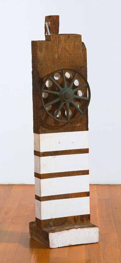 A sculpture consisting of a wooden beam with a haunched tenon on a wooden base. An iron wheel is affixed to the upper half of the front of the work; in between each spoke is a white painted orb. Four white stripes are painted around the bottom half of the sculpture, and the front of the base is also painted white.