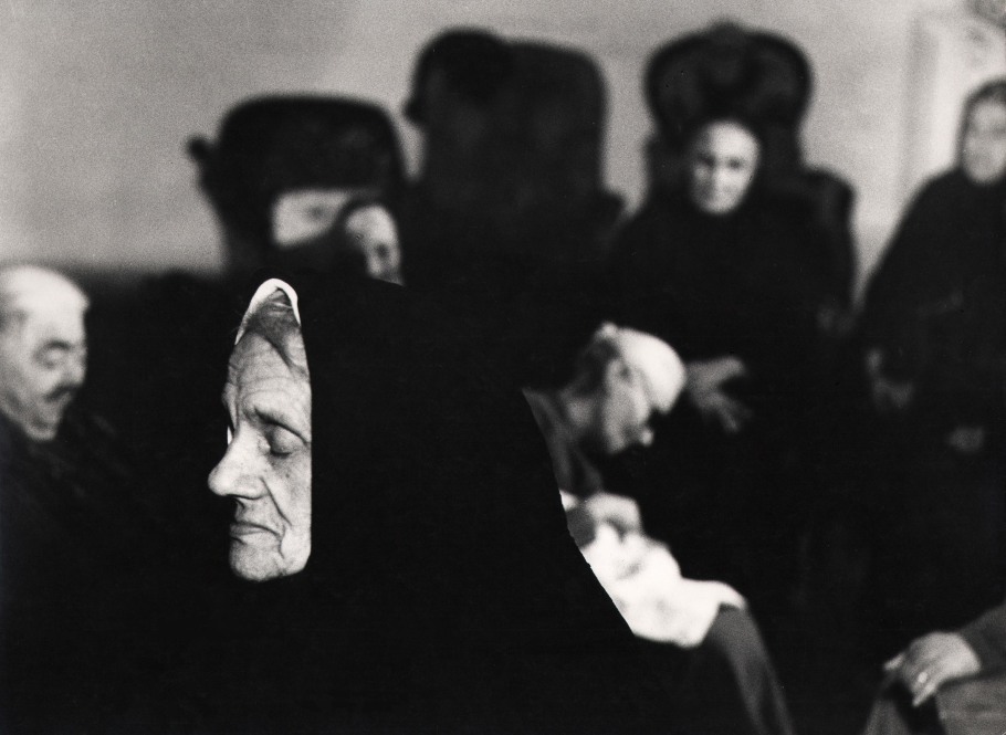 Mario Giacomelli, Verrà la morte e avrà i tuoi occhi, 1966–1968. High contrast image. A cloaked woman in the foreground with closed eyes, facing the left of the frame. Various figures out of focus behind her.