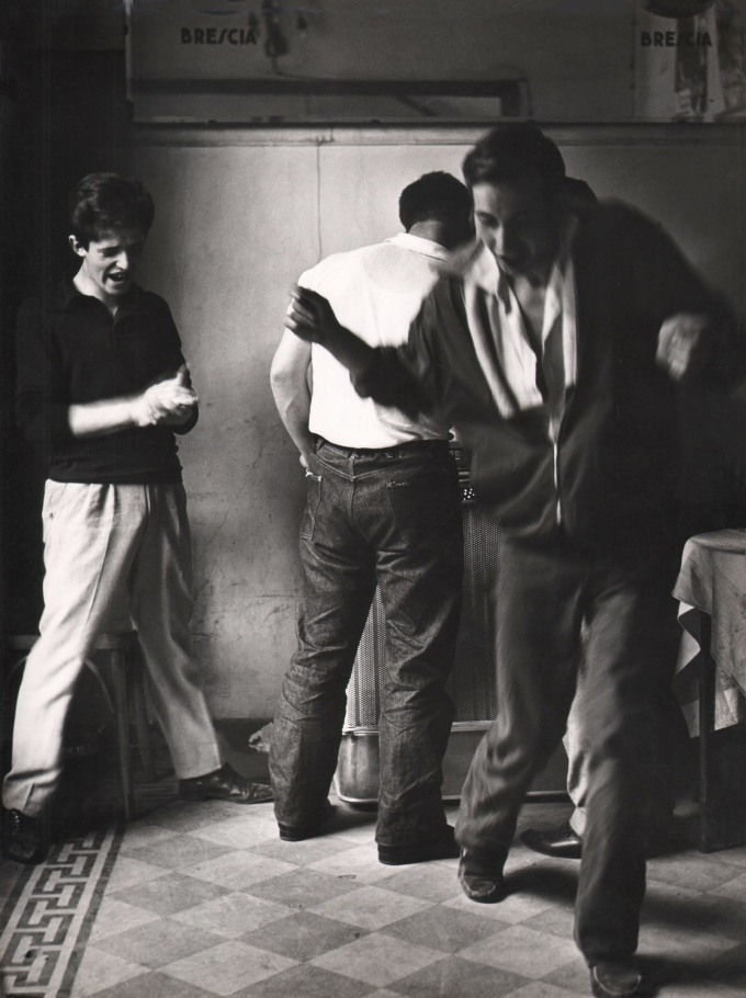 Mario Cattaneo, Jukebox, c. 1960. One man dances, blurred with motion, while another looks on clapping and a third man faces away from the camera towards the jukebox.