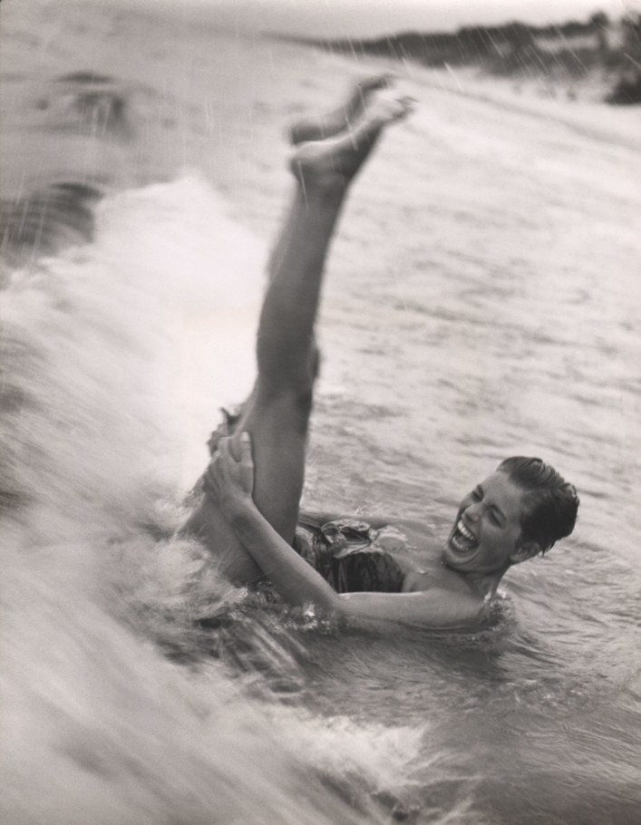Marvin D. Koehler, Untitled, ​c. 1955. A woman lays in shallow water amidst crashing waves, legs raised up above her. She is smiling and the scene is blurred with motion.