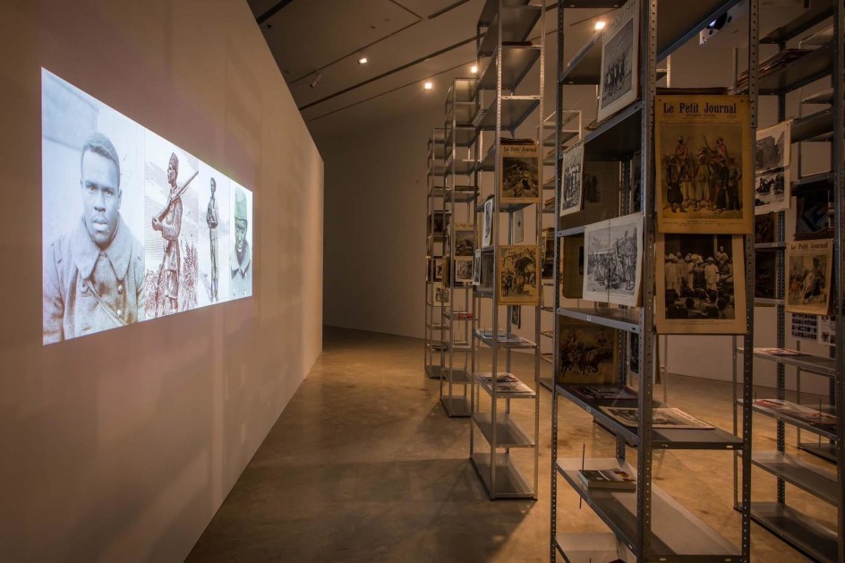  Kader Attia, installation view (left to right): The Debt, 2013, 2-channel digital video, projection, colour, based on the original 2-channel slide presentation; The Culture of Fear: An Invention of Evil, 2013, metal shelves, antique magazines, books, metal screws; Kader Attia, Museum of Contemporary Art Australia, Sydney, 2017, courtesy the artist, Galerie Nagel Draxler, Berlin/Cologne, Lehmann Maupin, New York and Hong Kong and Galleria Continua, San Gimignano, Les Moulins, Beijing, Habana. Image courtesy the artist and Museum of Contemporary Art Australia &copy; the artist, photograph: Anna Kučera